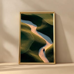 May include: A framed print depicting a golf course from above. A sandy path meanders through undulating green terrain. A golf cart is seen on the path. The frame is gold.