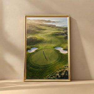 May include: A framed photograph of a golf course with a person standing on the green. The course is surrounded by green grass, sand traps, and the ocean in the background. The frame is gold.