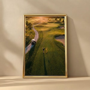May include: A framed print of a golf course at sunset. The image shows a golfer mid-swing on a green fairway, with a golf cart on a path. The scene is bathed in warm sunlight, with shadows cast across the course.