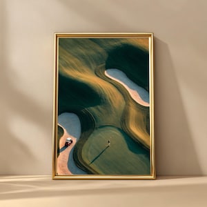 Puede incluir: Impresión enmarcada de un campo de golf, con un golfista, un carrito de golf y trampas de arena. El campo tiene una combinación de colores verde y beige, con largas sombras. El marco es dorado.