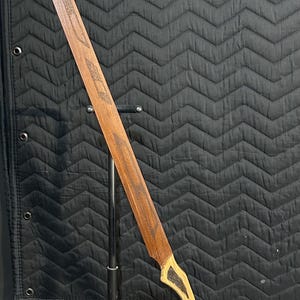 May include: A wooden sword with a curved blade and a light-colored handle. The blade is a dark brown color with etched designs. The handle has a contrasting light wood color with dark accents. The sword is displayed on a black metal stand.