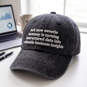 May include: A dark gray baseball cap with the text "not now sweetie mommy is turning unstructured data into actionable business insights" embroidered in white. The cap has a curved brim and a button on top.