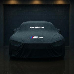 Puede incluir: Un coche oscuro cubierto con una funda protectora, estacionado en un garaje. La funda tiene el texto "KING SLEEPING" y el logotipo "M Power". La escena está mojada, con lluvia cayendo y reflejándose en el suelo.
