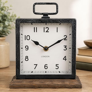 May include: A black square clock with a white face, black numerals, and black hands. The clock has a handle at the top and sits on a wooden base. The word "LONDON" is printed on the clock face.