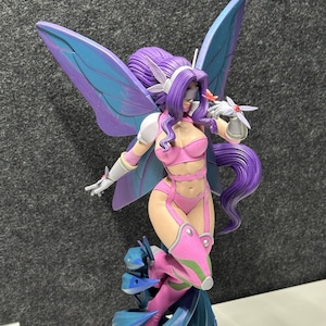 Peut inclure: Figurine détaillée d'un personnage fantastique avec des cheveux, des ailes et une tenue violets. La figurine est posée de manière dynamique, avec une palette de couleurs rose et violet. La base est de forme géométrique, et le fond est gris neutre.