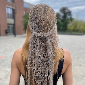 Può includere: Un copricapo realizzato in una rete metallica, drappeggiato sulla testa e che scende sulla schiena. La rete è un mix di tonalità argento, oro e bronzo, creando un effetto scintillante. Il copricapo è annodato sulla nuca.