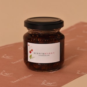May include: A jar of handmade jam with a black lid and a white label that reads "BERRY&MARRY". The jam is a dark reddish-brown colour and is sitting on pink paper with a repeating pattern. The label has a small illustration of berries.