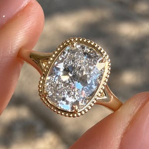 Puede incluir: Un anillo de oro con una gran piedra preciosa transparente de corte cojín rectangular engastada en un bisel de metal con cuentas. La banda es de oro amarillo pulido y presenta un diseño de vástago dividido que conecta al engaste.