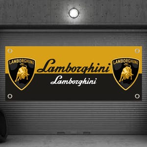 Puede incluir: Una pancarta con el logotipo de Lamborghini, en negro y dorado. La pancarta presenta el nombre Lamborghini en una fuente estilizada. El fondo es un entorno de garaje con neumáticos y una puerta metálica.