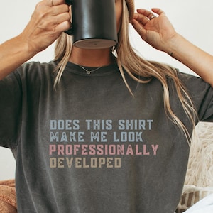 Peut inclure: Un t-shirt gris anthracite à manches courtes avec le texte "DOES THIS SHIRT MAKE ME LOOK PROFESSIONALLY DEVELOPED" imprimé en lettres capitales bleu clair, rose et beige. Le vêtement présente une coupe décontractée et un aspect délavé.