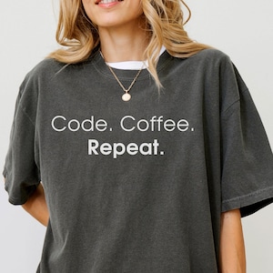 May include: A charcoal grey short-sleeve t-shirt featuring the white text Code. Coffee. Repeat. printed on the chest. The garment has a crew neckline and a relaxed, loose-fitting silhouette with a slightly faded, washed-out fabric appearance.
