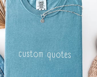 Peut inclure: Un t-shirt bleu à manches courtes avec le texte "custom quotes" imprimé en blanc sur le devant. Le t-shirt présente une étiquette de marque Comfort Colors à l'encolure intérieure et est accompagné d'un collier à chaîne argentée avec un pendentif.