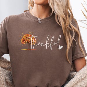 May include: A brown short-sleeve t-shirt featuring a graphic of an autumn tree with orange and yellow leaves. The word "thankful" is printed in white cursive script next to the tree, accompanied by a small white heart icon on the right side.