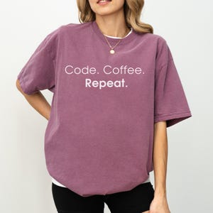 May include: A plum-coloured short-sleeve t-shirt featuring the white text Code. Coffee. Repeat. printed on the chest. The garment has a crew neckline and a relaxed fit, paired with a simple gold-toned chain necklace worn around the neck.