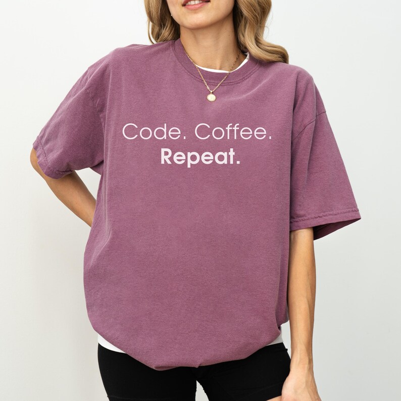 May include: A plum-coloured short-sleeve t-shirt featuring the white text Code. Coffee. Repeat. printed on the chest. The garment has a crew neckline and a relaxed fit, paired with a simple gold-toned chain necklace worn around the neck.