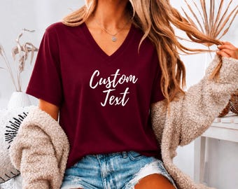Peut inclure: Un t-shirt bordeaux à col en V avec le texte Custom Text écrit en blanc sur le devant. Le t-shirt est porté avec un gilet texturé beige et un short en jean bleu. Le vêtement possède des manches courtes et une coupe standard décontractée.