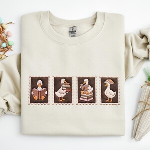 Pode incluir: Uma camisola de gola redonda cor de areia com quatro emblemas retangulares bordados, cada um representando um pato branco a interagir com livros. Os desenhos mostram um pato a ler, a carregar uma pilha de livros, sentado sobre livros e com um livro na cabeça.