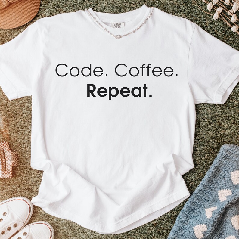 May include: A white short-sleeved t-shirt featuring the black text Code. Coffee. Repeat. printed on the front. The shirt has a crew neckline and a visible brand label on the inside collar area, presented in a clean and simple design.