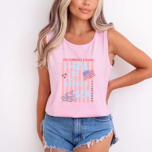 May include: A light pink sleeveless tank top featuring a graphic print with vertical stripes and the text 250 SUMMERS STRONG FROM SEA TO SHINING SEA 1776 2026. The design includes a small American flag, stars, and a stylized ocean wave graphic.