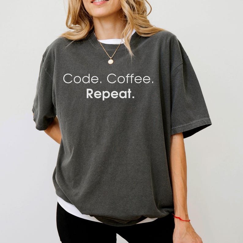 May include: A charcoal grey short-sleeve t-shirt featuring the white text Code. Coffee. Repeat. printed on the chest. The garment has a crew neckline and a relaxed, loose-fitting silhouette with a slightly faded, washed-out fabric appearance.