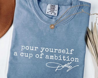 Peut inclure: Un t-shirt à manches courtes de couleur bleu jean avec une encolure ronde. Le vêtement présente le texte imprimé en blanc "pour yourself a cup of ambition" suivi de la signature "Dolly" en écriture cursive blanche. Un collier doré est posé.