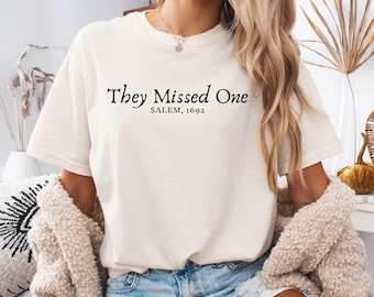 May include: A cream-coloured short-sleeve t-shirt featuring black text printed on the chest that reads "They Missed One" in a serif font, with "SALEM, 1692" printed in a smaller sans-serif font directly underneath the main phrase.