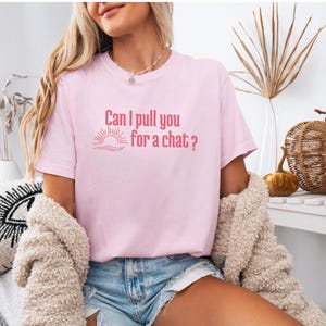 May include: A light pink short-sleeved t-shirt featuring the dark pink text Can I pull you for a chat? accompanied by a graphic of a rising sun over waves. The shirt has a crew neckline and is worn with light blue denim shorts and a beige cardigan.