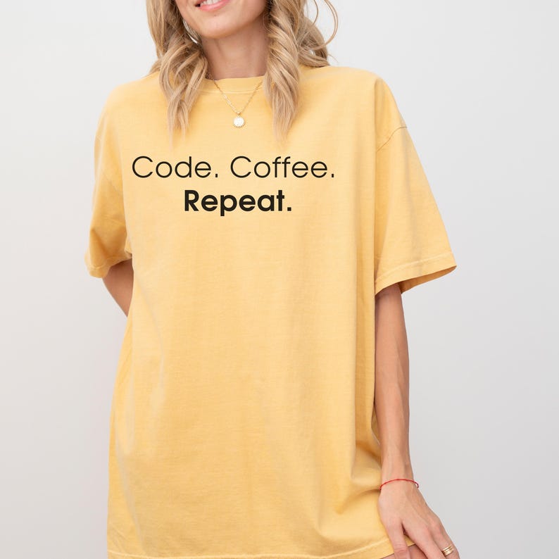 May include: A mustard yellow short-sleeve t-shirt featuring the black text Code. Coffee. Repeat. printed on the front. The garment has a crew neckline and a relaxed fit, showcasing a simple design with bold lettering across the chest area.