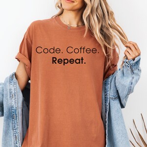 May include: A rust-coloured short-sleeve t-shirt featuring the black text Code. Coffee. Repeat. printed on the front. The shirt has a crew neckline and is worn with a light blue denim jacket draped over the shoulders.