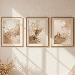 May include: Three framed abstract watercolor art prints in neutral tones of beige, brown, and gold. The artwork features flowing shapes and lines, with a minimalist aesthetic. The frames are a light wood color, and the overall style is modern and elegant.