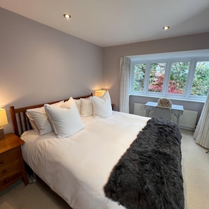 May include: A bedroom featuring a wooden bed frame with white bedding and pillows. A dark grey faux fur throw is placed at the foot of the bed. A wooden bedside table with a lamp is on the left. A window with a view is on the right.