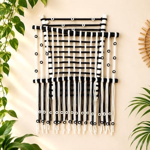 May include: A wall hanging featuring a grid of black and white cord woven over horizontal wooden dowels. The design includes small circular black and white beads integrated into the weave and hanging fringe at the bottom with beads.