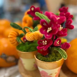 May include: Two small decorative flower pots with artificial flowers. One pot has bright pink flowers, the other has orange flowers. The pots are cream-colored with gold trim and sit on a wooden tray. The flowers are made of a soft, fuzzy material.