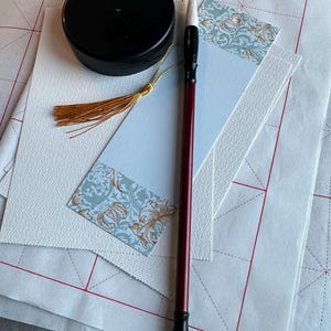 May include: A calligraphy set featuring a dark red and black brush with a white tip, a black circular ink container, and a light blue bookmark with a gold tassel and floral pattern, all resting on white paper sheets with a red grid pattern.