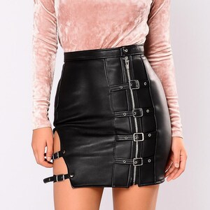 May include: A long-sleeved, blush pink velvet top paired with a black faux leather mini skirt. The skirt features a zipper closure and multiple buckle details, with a slit on the side.