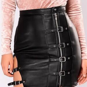 May include: Black faux leather skirt with a high waist and a zipper down the front. The skirt features silver buckles and straps on the front and side, and a slit on the side. The skirt is worn with a pink long-sleeve top.