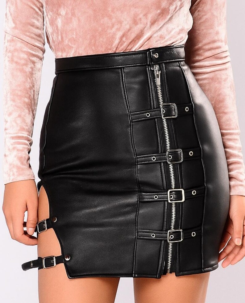 May include: Black faux leather skirt with a high waist and a zipper down the front. The skirt features silver buckles and straps on the front and side, and a slit on the side. The skirt is worn with a pink long-sleeve top.