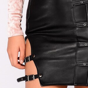 May include: Black faux leather skirt with side slits and silver buckle details. The skirt features a vertical seam with decorative silver rivets and a zipper. The garment is a stylish piece of apparel.