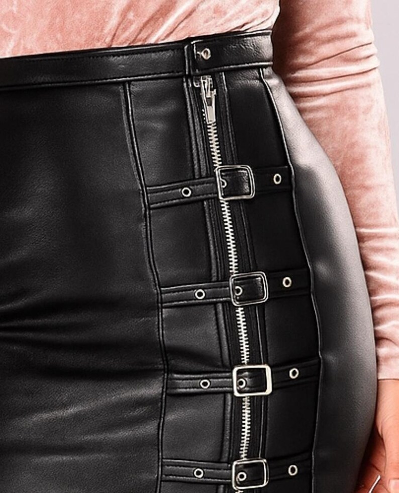 May include: Black faux leather skirt with a zipper closure and buckle details. The skirt features a vertical zipper running down the front and decorative straps with silver buckles and eyelets. The skirt is a pencil skirt style.