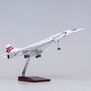 Può includere: Modello bianco di un aereo Concorde, con la livrea British Airways. Il modello è montato su un supporto argentato con una base in legno scuro. L'aereo ha un design alare a delta distintivo e un lungo muso appuntito.