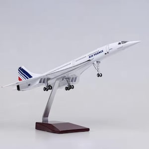 Può includere: Un modello bianco di aereo Concorde Air France con dettagli blu e rossi sulla coda. Il modello è montato su un supporto argentato con una base marrone scuro. La scritta "AIR FRANCE" è stampata sul lato.