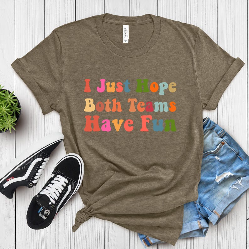 Peut inclure: Un t-shirt vert olive chin&eacute; avec le texte "I Just Hope Both Teams Have Fun" dans une police r&eacute;tro color&eacute;e. Le t-shirt est associ&eacute; &agrave; un short en jean et &agrave; des baskets noires et blanches.