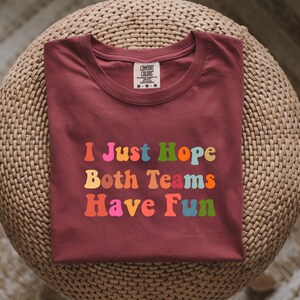 Peut inclure: Un t-shirt Comfort Colors bordeaux avec le texte "I Just Hope Both Teams Have Fun" dans des lettres color&eacute;es de style r&eacute;tro. Le t-shirt est pli&eacute; et pos&eacute; sur une surface circulaire tiss&eacute;e.