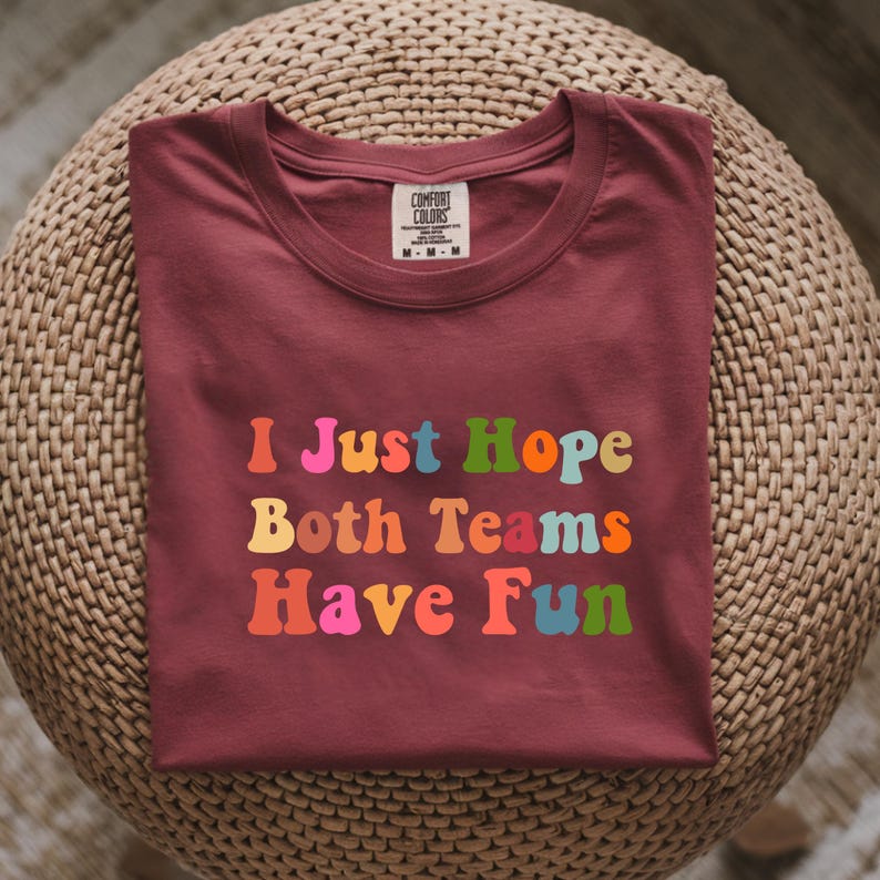 Peut inclure: Un t-shirt Comfort Colors bordeaux avec le texte "I Just Hope Both Teams Have Fun" dans des lettres color&eacute;es de style r&eacute;tro. Le t-shirt est pli&eacute; et pos&eacute; sur une surface circulaire tiss&eacute;e.