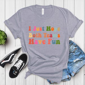 Peut inclure: T-shirt gris clair avec l'inscription "I Just Hope Both Teams Have Fun" dans des lettres color&eacute;es de style r&eacute;tro. Le t-shirt est associ&eacute; &agrave; des baskets noires et blanches et &agrave; un short en jean.