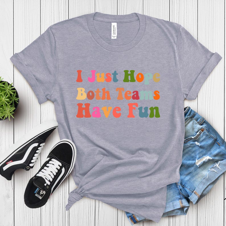 Peut inclure: T-shirt gris clair avec l'inscription "I Just Hope Both Teams Have Fun" dans des lettres color&eacute;es de style r&eacute;tro. Le t-shirt est associ&eacute; &agrave; des baskets noires et blanches et &agrave; un short en jean.