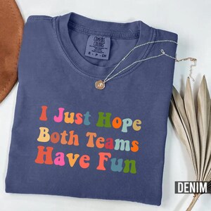 Peut inclure: Un t-shirt bleu denim pli&eacute; avec l'inscription "I Just Hope Both Teams Have Fun" en lettres color&eacute;es. Un collier argent&eacute; avec un pendentif est pos&eacute; sur le t-shirt. Le t-shirt est pr&eacute;sent&eacute; sur une surface neutre.