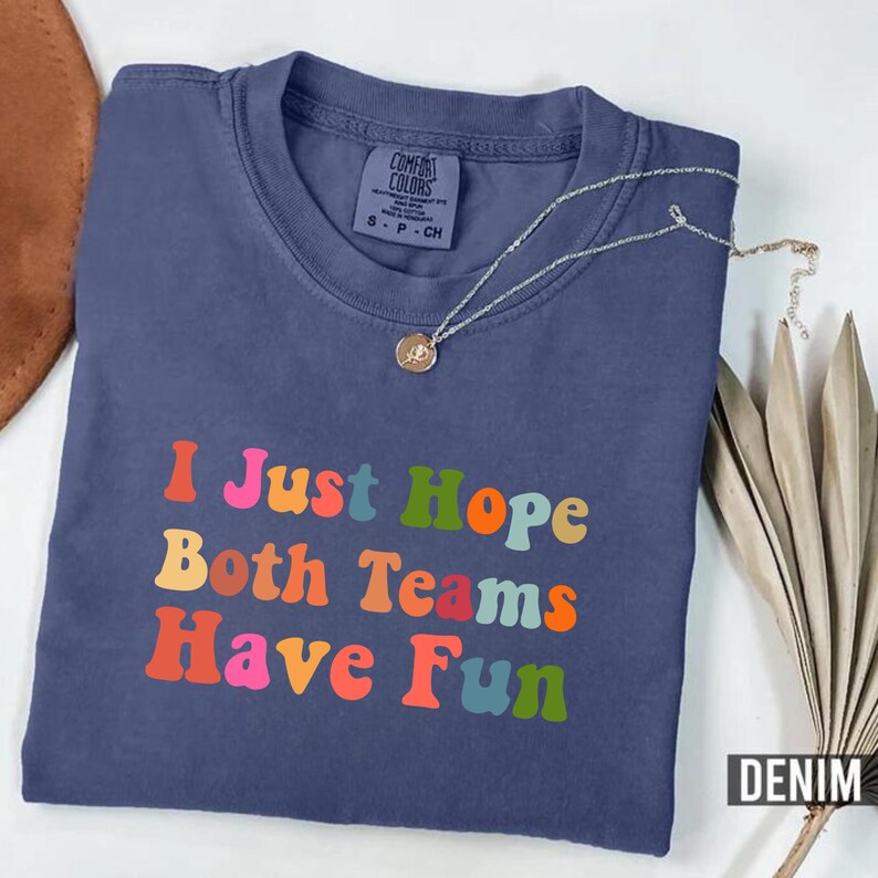 Peut inclure: Un t-shirt bleu denim pli&eacute; avec l'inscription "I Just Hope Both Teams Have Fun" en lettres color&eacute;es. Un collier argent&eacute; avec un pendentif est pos&eacute; sur le t-shirt. Le t-shirt est pr&eacute;sent&eacute; sur une surface neutre.