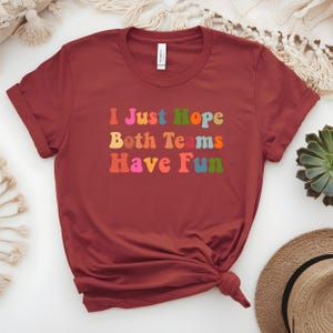 Peut inclure: Un t-shirt bordeaux &agrave; manches retrouss&eacute;es. Le t-shirt affiche le texte "I Just Hope Both Teams Have Fun" dans des lettres color&eacute;es de style r&eacute;tro. Le t-shirt est nou&eacute; en bas.