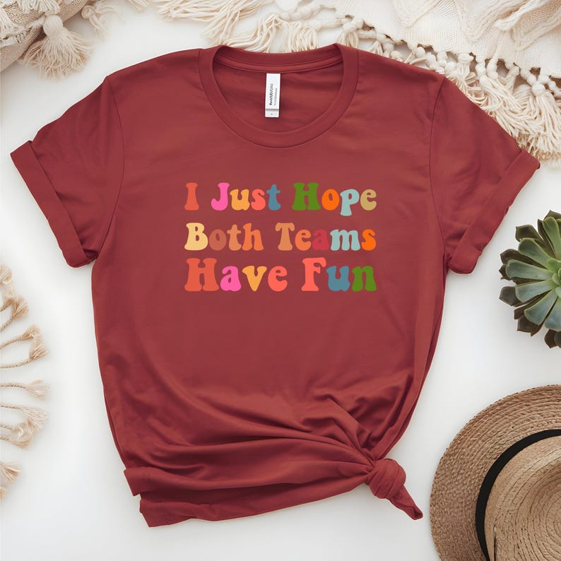 Peut inclure: Un t-shirt bordeaux &agrave; manches retrouss&eacute;es. Le t-shirt affiche le texte "I Just Hope Both Teams Have Fun" dans des lettres color&eacute;es de style r&eacute;tro. Le t-shirt est nou&eacute; en bas.